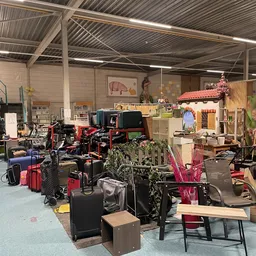 Overzicht van de kringloopwinkel met kledingrekken, koffers, huishoudspullen en meubels verspreid over de vloer