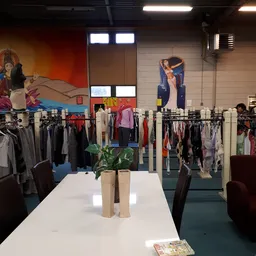 Winkelvloer van kringloopwinkel met kledingrekken en muurschilderingen, witte tafel met plantenbakken in voorgrond