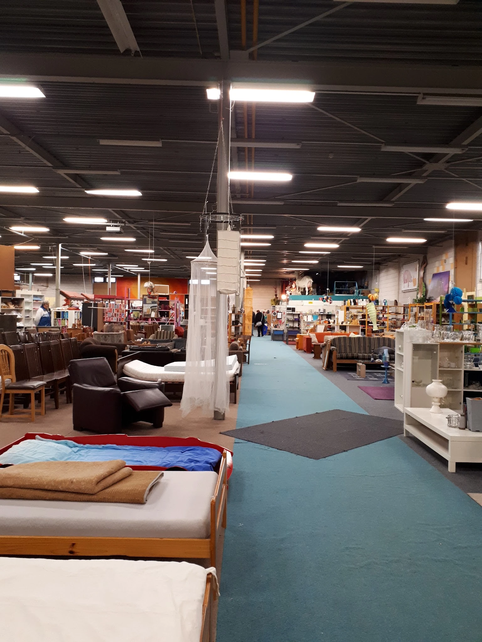Kringloopwinkel met blauwe vloer, bruine stoelen, stellingen met spullen en verlichte plafondlampen
