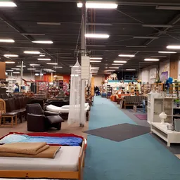 Kringloopwinkel met blauwe vloer, bruine stoelen, stellingen met spullen en verlichte plafondlampen