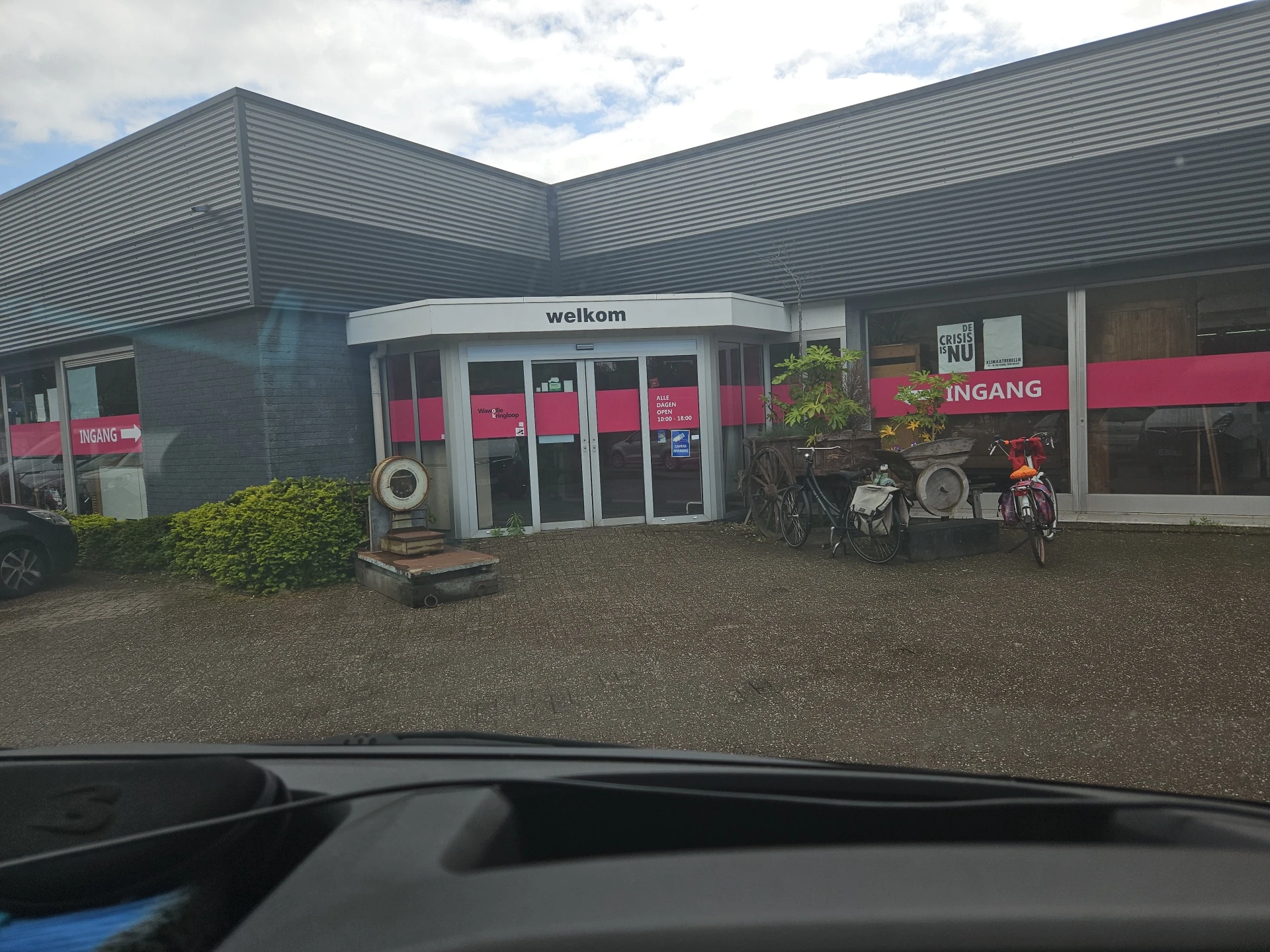 Grijze kringloopwinkel met roze accenten, welkom-bord en ingang zichtbaar, fietsen geparkeerd.