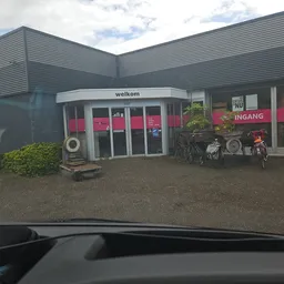Grijze kringloopwinkel met roze accenten, welkom-bord en ingang zichtbaar, fietsen geparkeerd.
