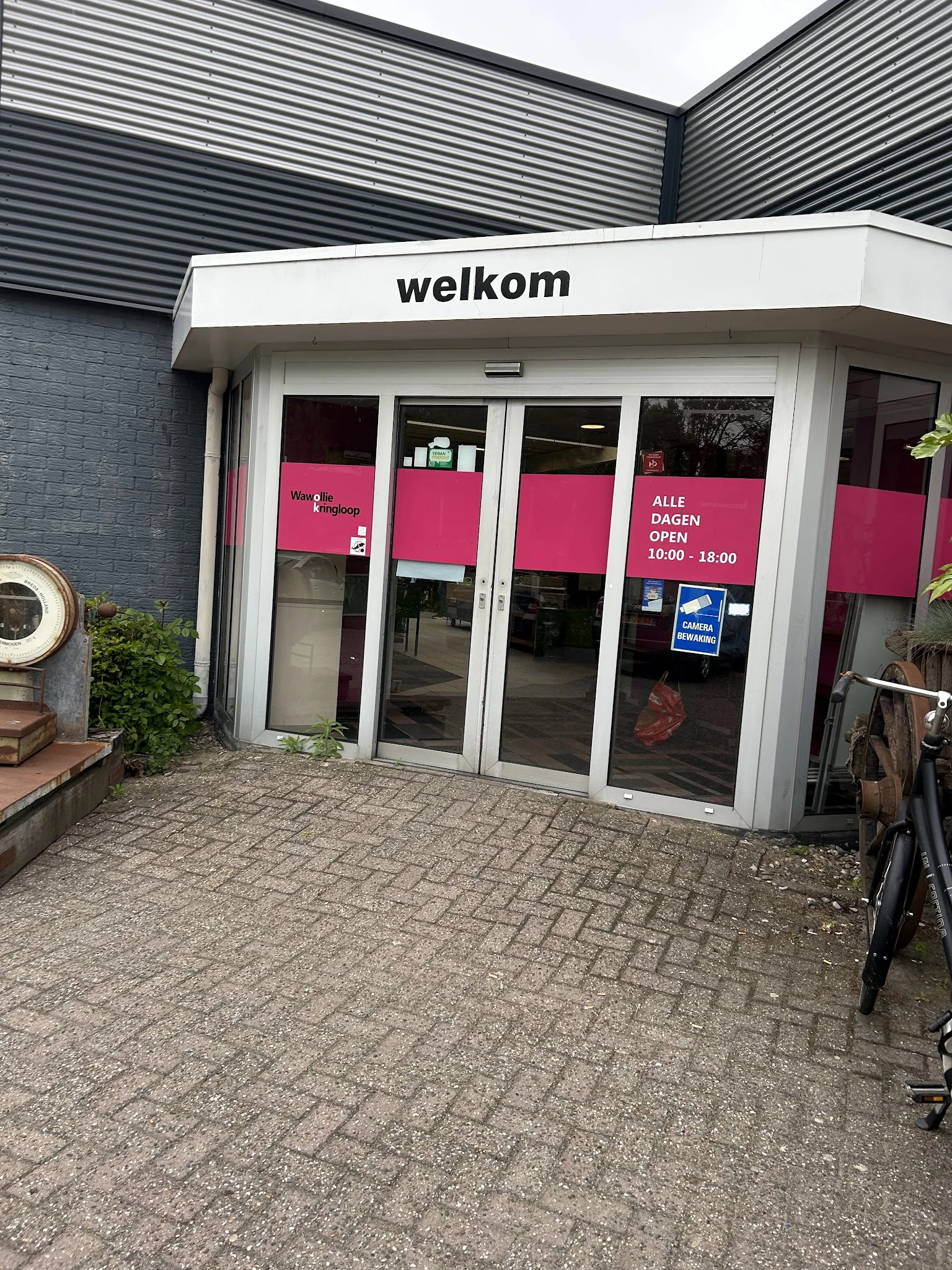 Gevel van kringloopwinkel met witte aluminium pui, roze belettering en openingstijden op raam