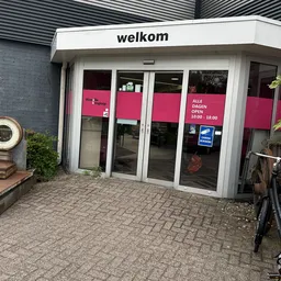 Gevel van kringloopwinkel met witte aluminium pui, roze belettering en openingstijden op raam