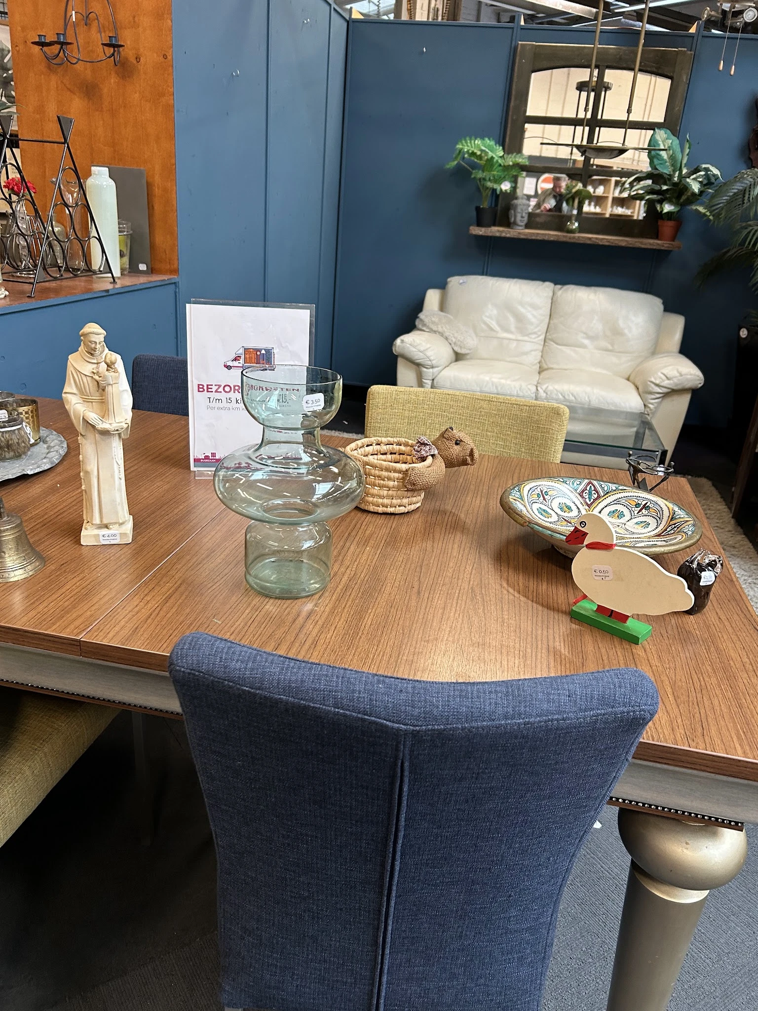 Woonkamer van kringloopwinkel met houten tafel, decoratieve items, glazen vaas, borden en crème-kleurige bank tegen blauwe mu