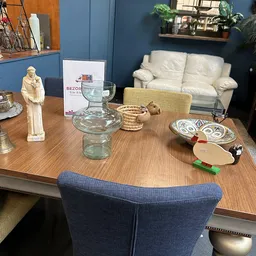 Woonkamer van kringloopwinkel met houten tafel, decoratieve items, glazen vaas, borden en crème-kleurige bank tegen blauwe mu