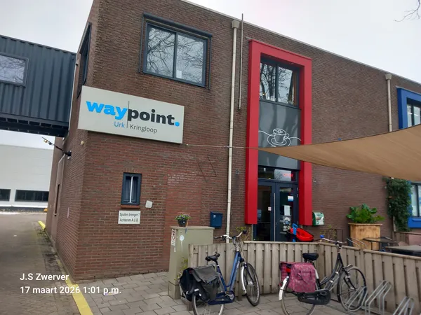 Bakstenen gevel van kringloopwinkel Waypoint met blauwe logo, rode deur en ingang met fietsen ervoor