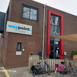 Bakstenen gevel van kringloopwinkel Waypoint met blauwe logo, rode deur en ingang met fietsen ervoor