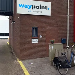Bakstenen gevel van kringloopwinkel met raam, deur en blauwe tropische plant, fiets en afvalbak op straat
