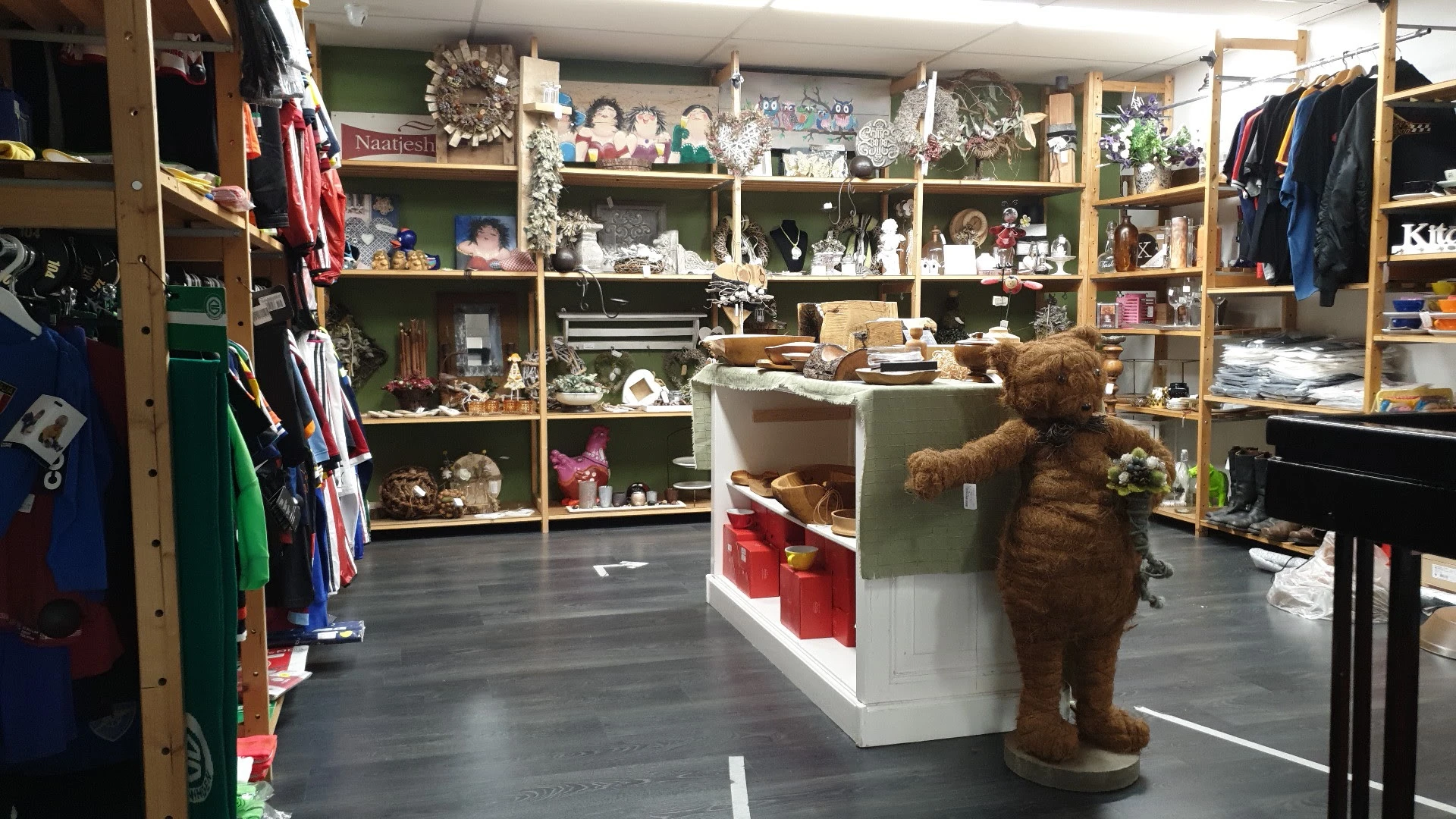 Winkelinterieur met houten stellingkasten tegen groene muren, gevuld met huishoudartikelen, decoratie en serviesgoed. Bruine 