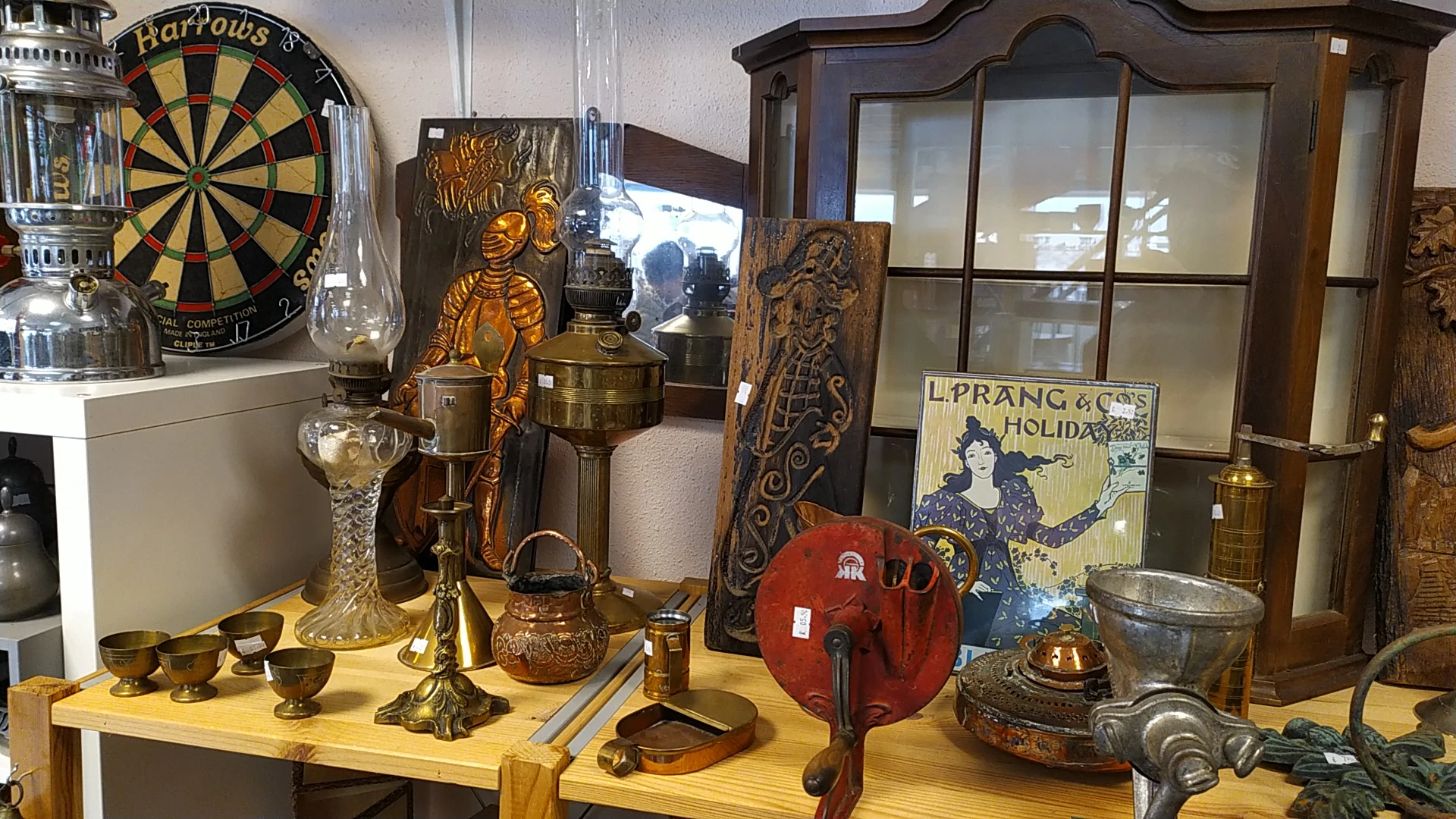 Vitrinekast met antieke en vintage objecten: gouden beeldjes, koperen lantaarns, glazen karaf, rode handschoenen, dartbord en