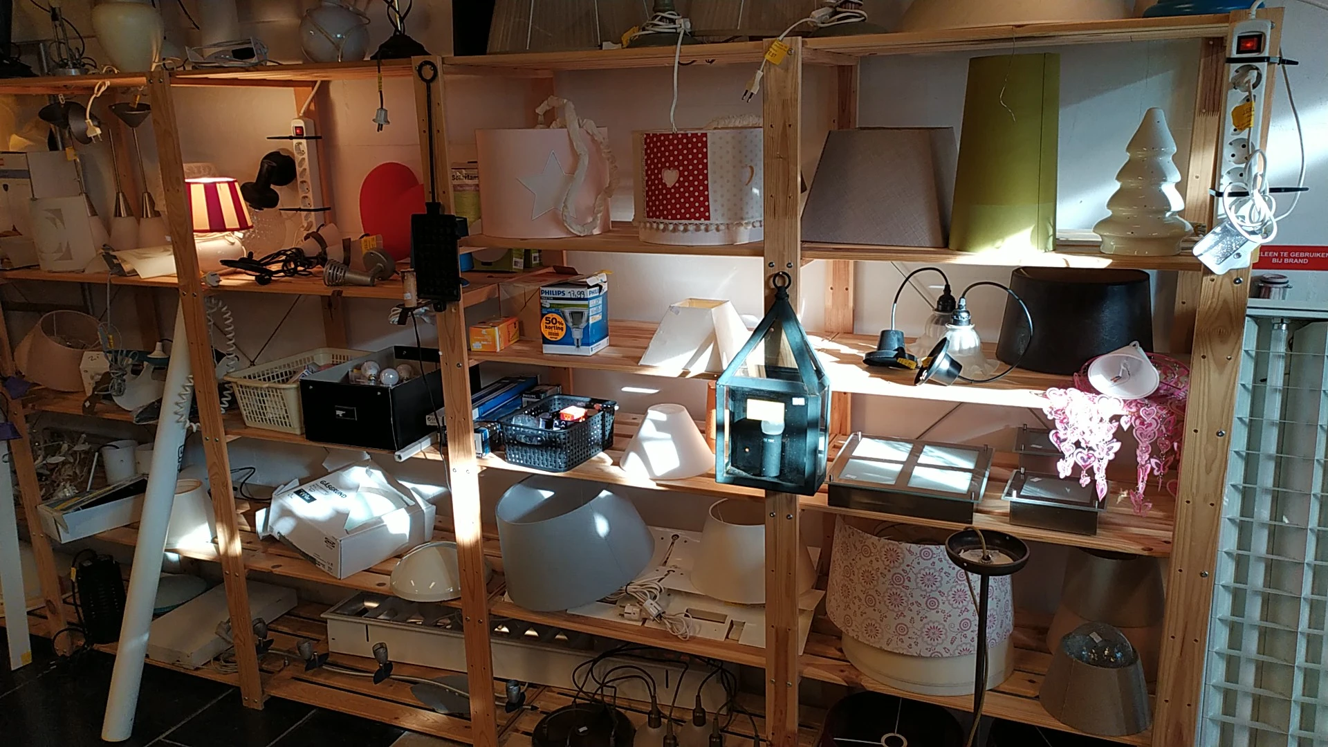 Houten stellingsysteem met diverse items zoals lampen, dozen, stoelen en huishoudelijke spullen in een kringloopwinkel