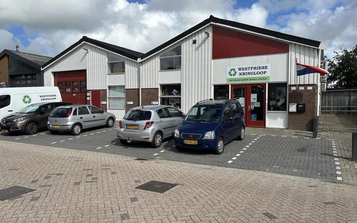 Witte kringloopwinkel met rode accenten, parkeerplaats met auto's ervoor, helder daglicht