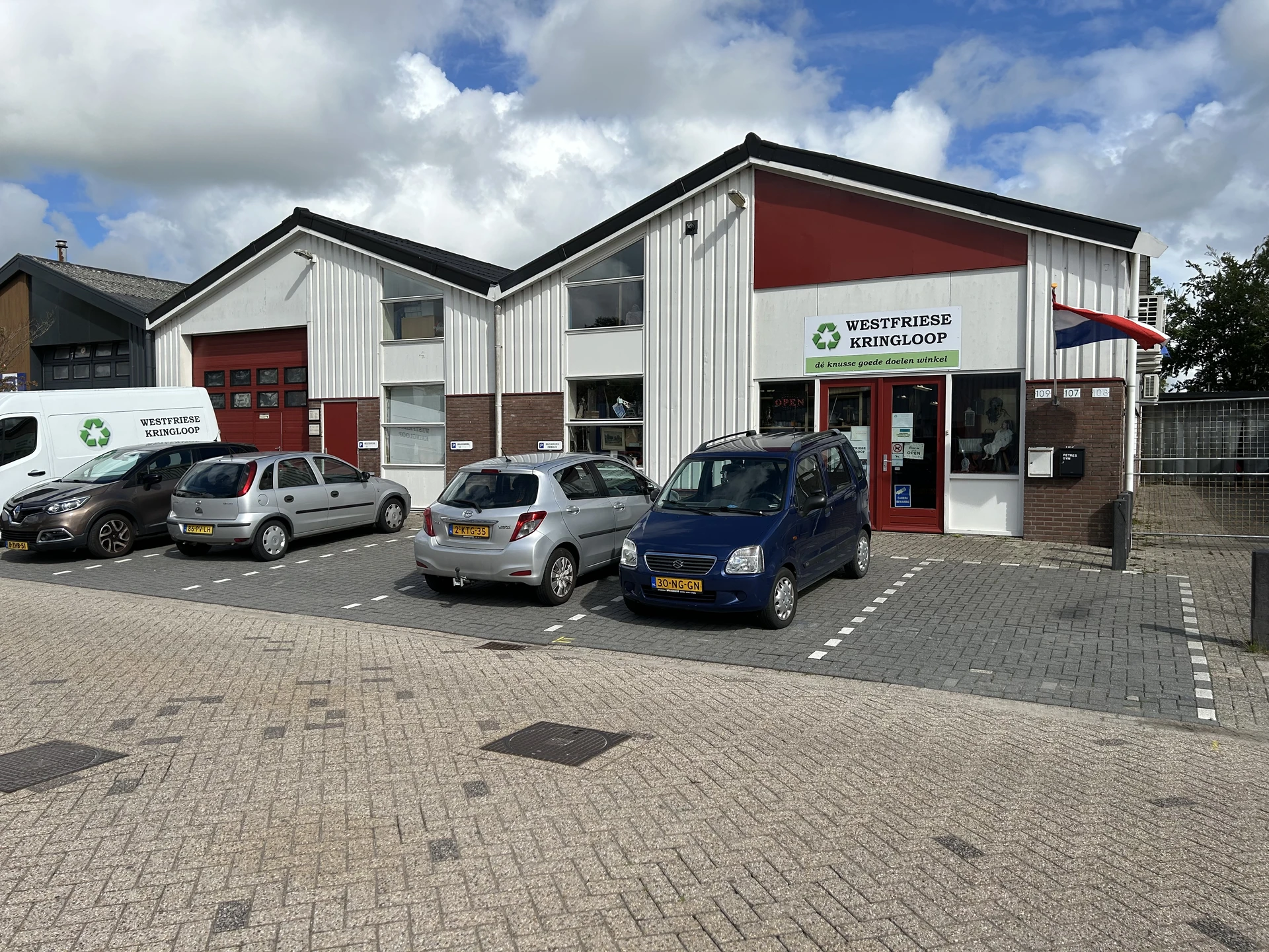 Witte kringloopwinkel met rode accenten, parkeerplaats met auto's ervoor, helder daglicht