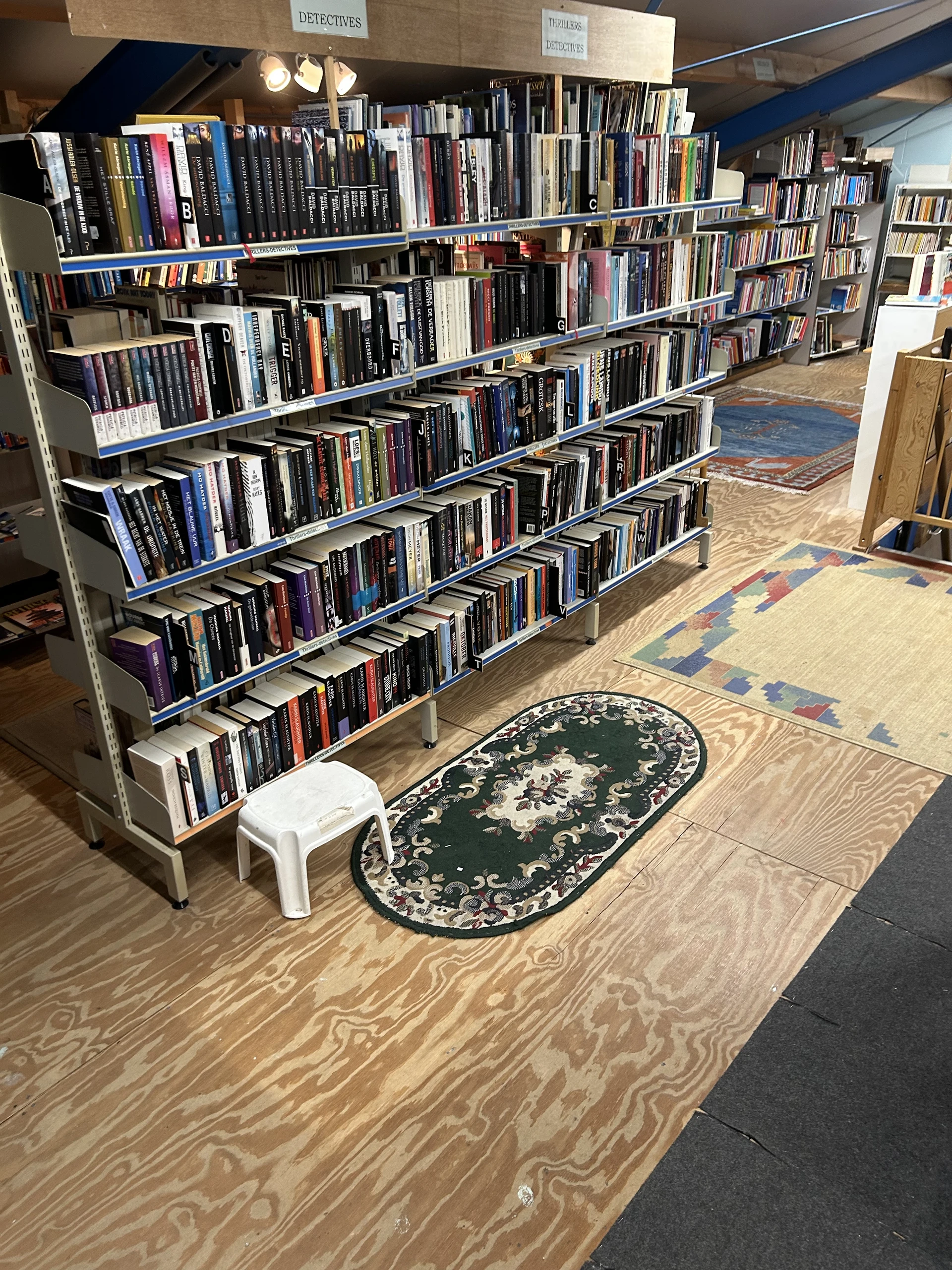 Tweedehands cd's en dvd's op lange stellingen in kringloopwinkel, met groene oval vloerkleed en witte krukje op houten vloer