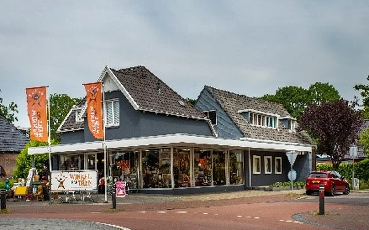 Donkerblauwe kringloopwinkel met witte afdak en oranje banners aan de voorkant, aan een plein met rode bestrating