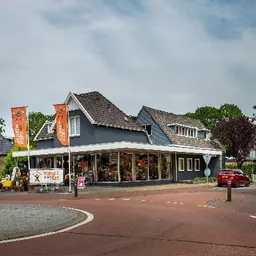 Donkerblauwe kringloopwinkel met witte afdak en oranje banners aan de voorkant, aan een plein met rode bestrating