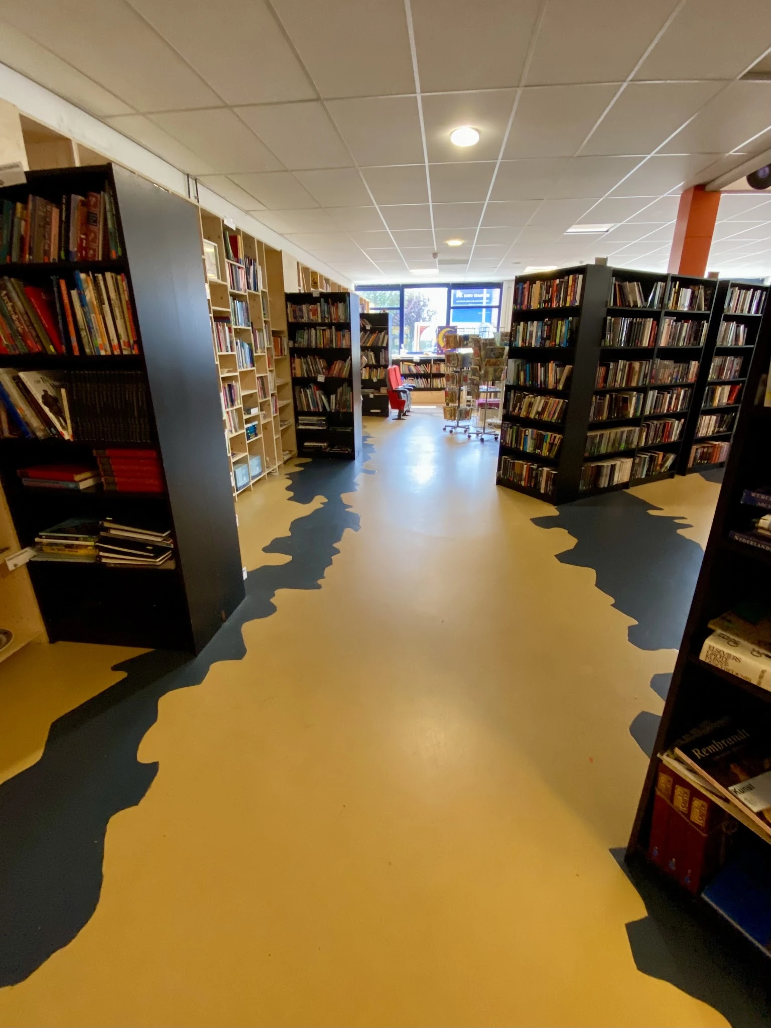 Binnenaanzicht van kringloopwinkel met gele en donkergrijze vloer, boekenkasten langs de muren en boeken in stellingen