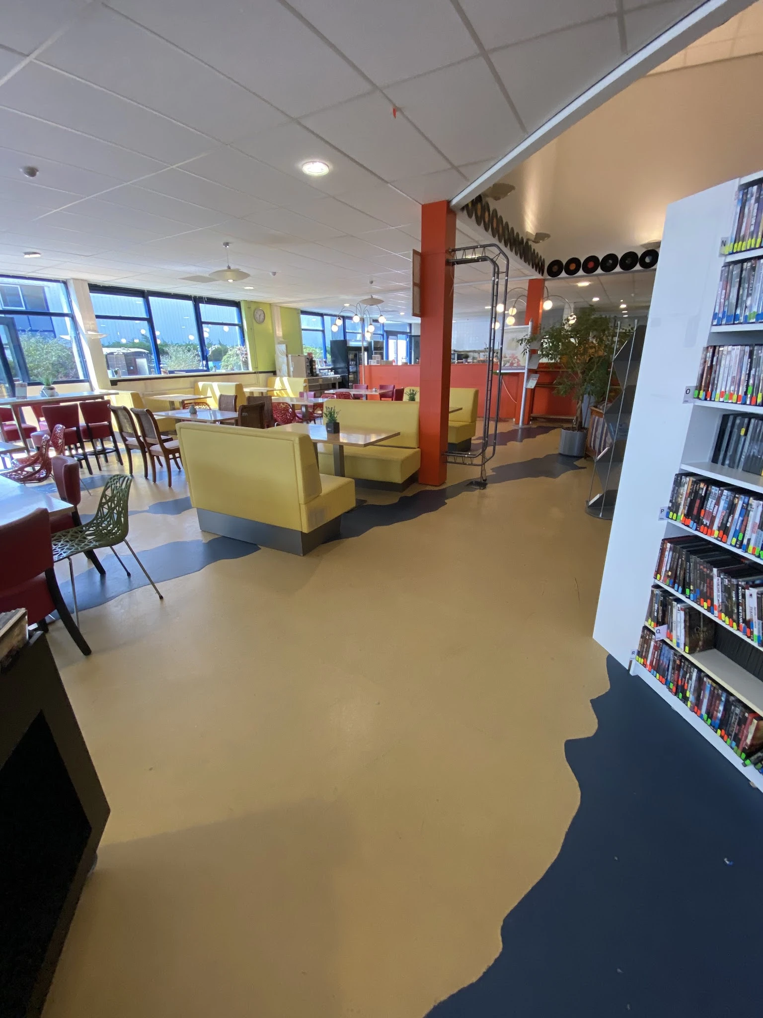 Winkelvloer met gele zithoeken, rode kolommen, boekenrekken en grote ramen met uitzicht