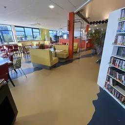 Winkelvloer met gele zithoeken, rode kolommen, boekenrekken en grote ramen met uitzicht