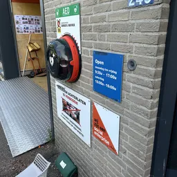 Voorkant kringloopwinkel met stenen gevel, openingstijden op blauw bord, rode en zwarte speelgoed-auto aan muur