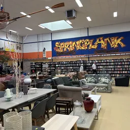 Ruime winkelruimte met boekenkasten, zithoeken met fauteuils en een groot blauw decoratief bord aan de wand