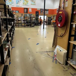 Winkelvloer van kringloopwinkel met stellingen, spullen aan meubelstukken, rode brandslangkast aan rechterkant en blauwe voet
