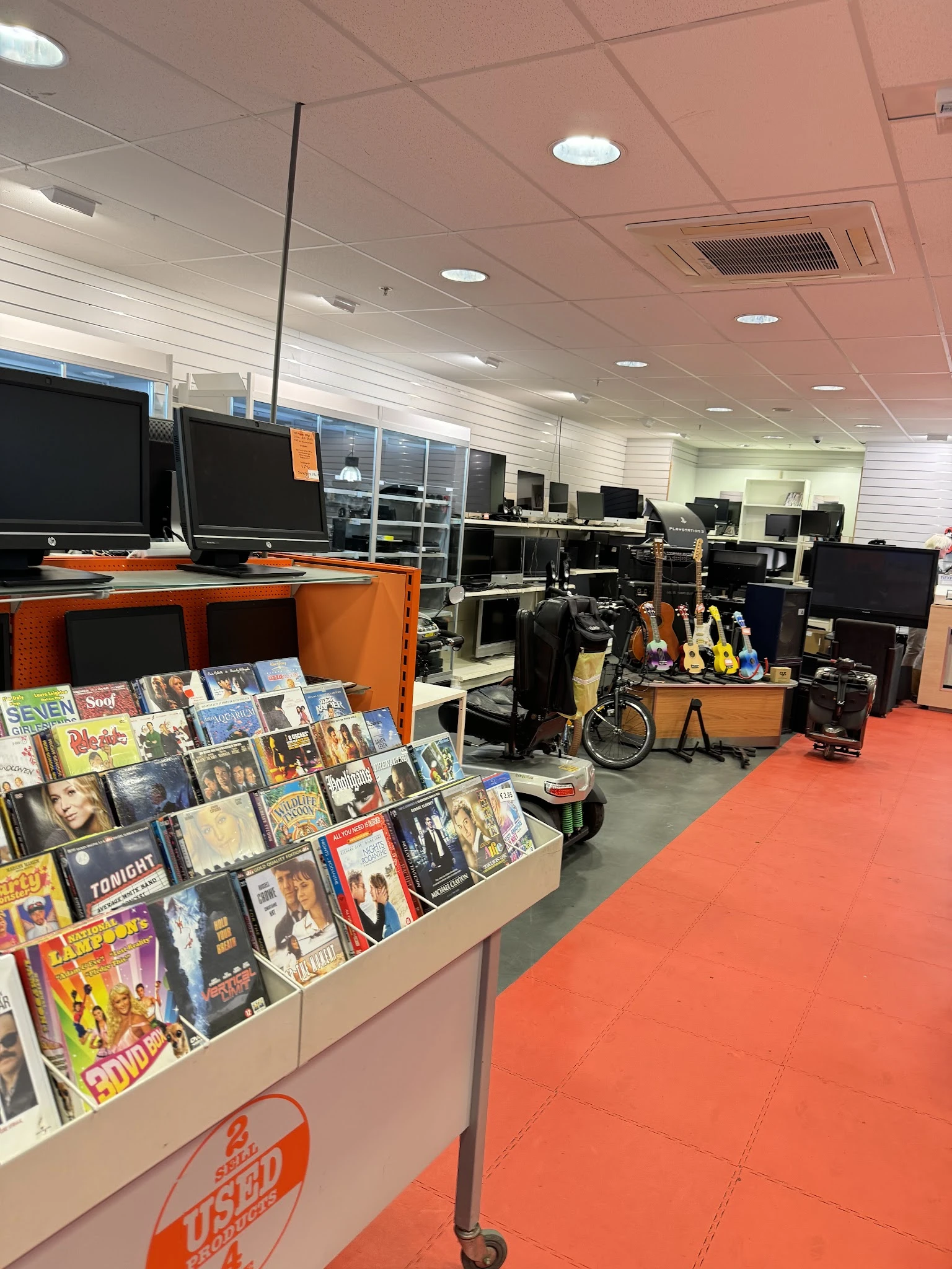 Kringloopwinkelinterieur met computermonitoren, oranje meubels, muziekinstrumenten en dvd's aan het raam