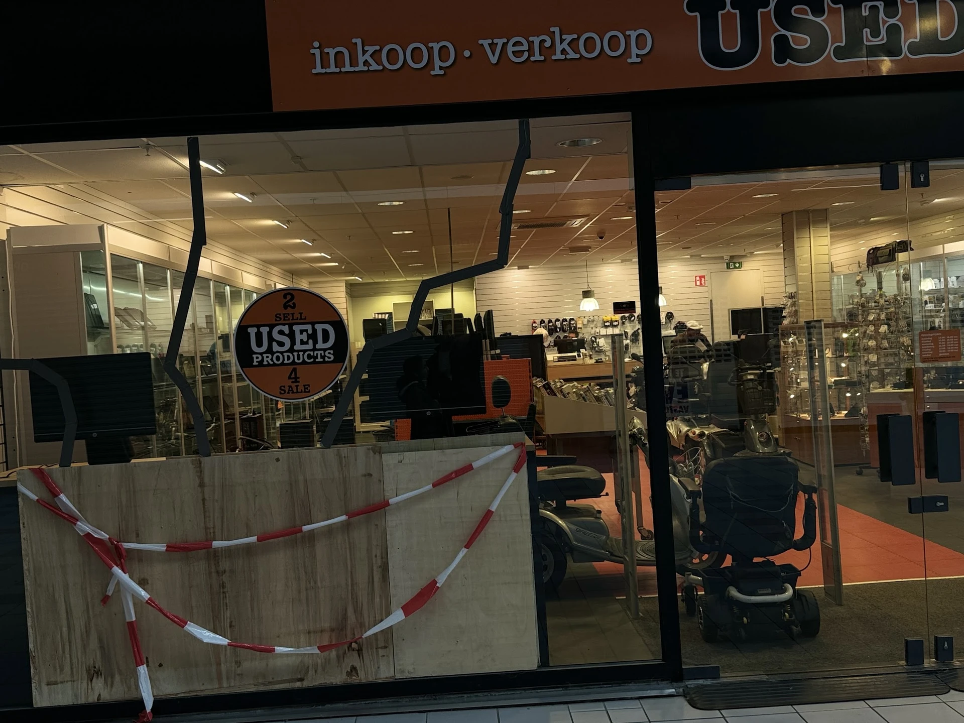 Winkelinterieur met gesloten deur, rood-wit lint, kinderwagens en diversen huishoudartikelen op rekken