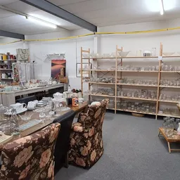 Binnenzijde kringloopwinkel met houten stellingen vol glazen potten, twee bruine bloemenpatroonstofels en werkplekken