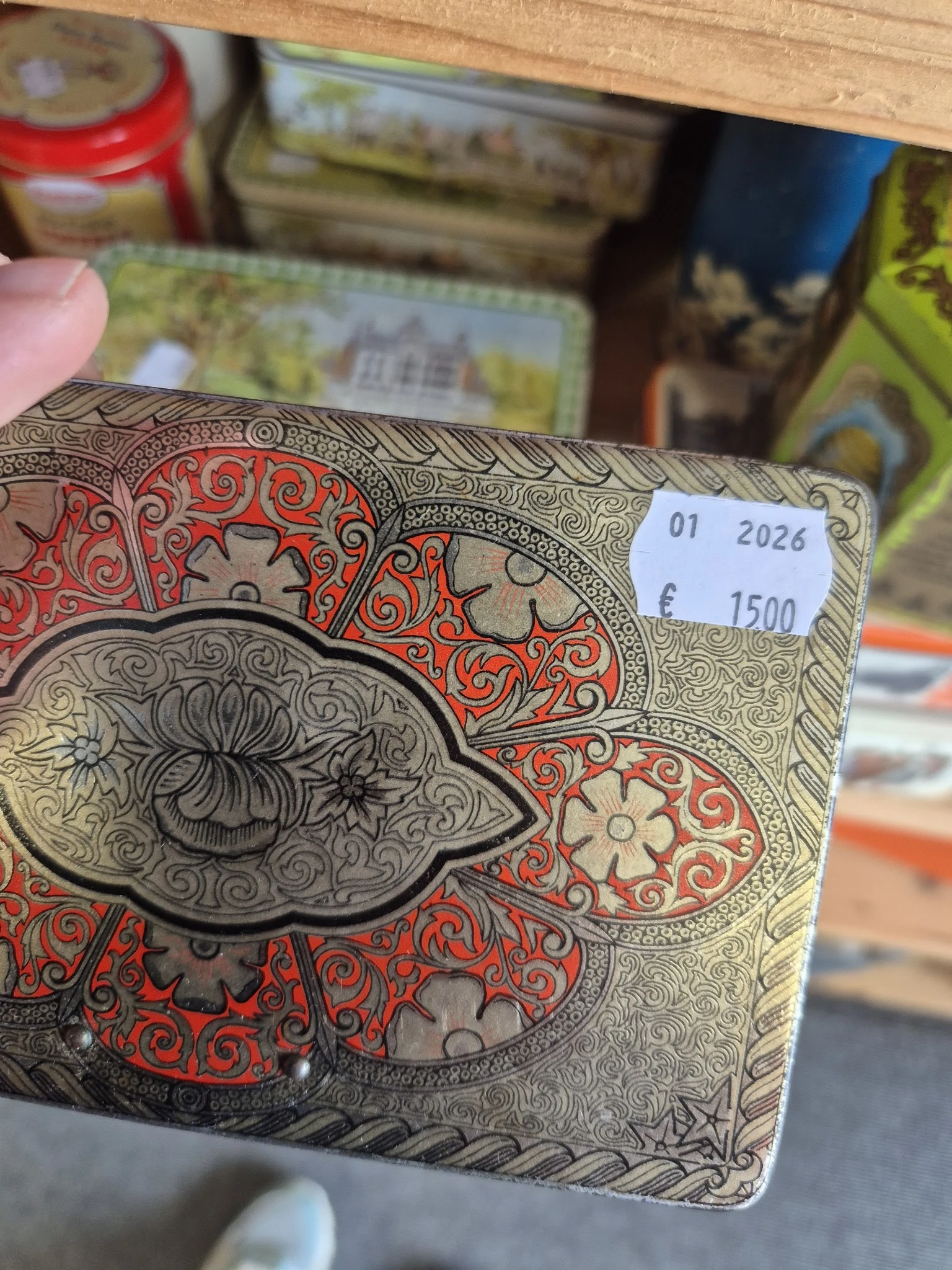 Close-up van een decoratief boek of object met rood en beige ornamentale patronen en een oog-motief, met een prijssticker