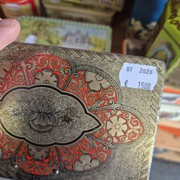 Close-up van een decoratief boek of object met rood en beige ornamentale patronen en een oog-motief, met een prijssticker