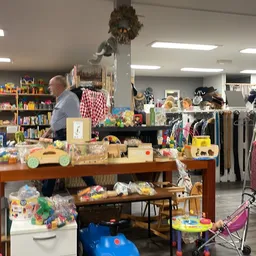 Winkelvloer van kringloopwinkel met tafels vol speelgoed, kleding aan rekken, boeken op achtergrond en klanten aan het winkel
