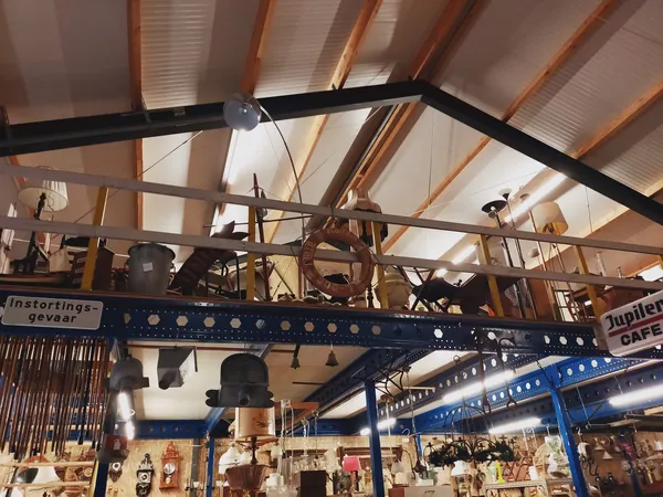 Zicht op het plafond en kranen van een kringloopwinkel met houten balken, metalen constructies en diverse spullen eronder