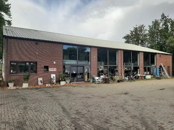 Voorgevel van een bakstenen kringloopwinkel met grote raampartijen en open deuren, omgeven door een parkeerterrein