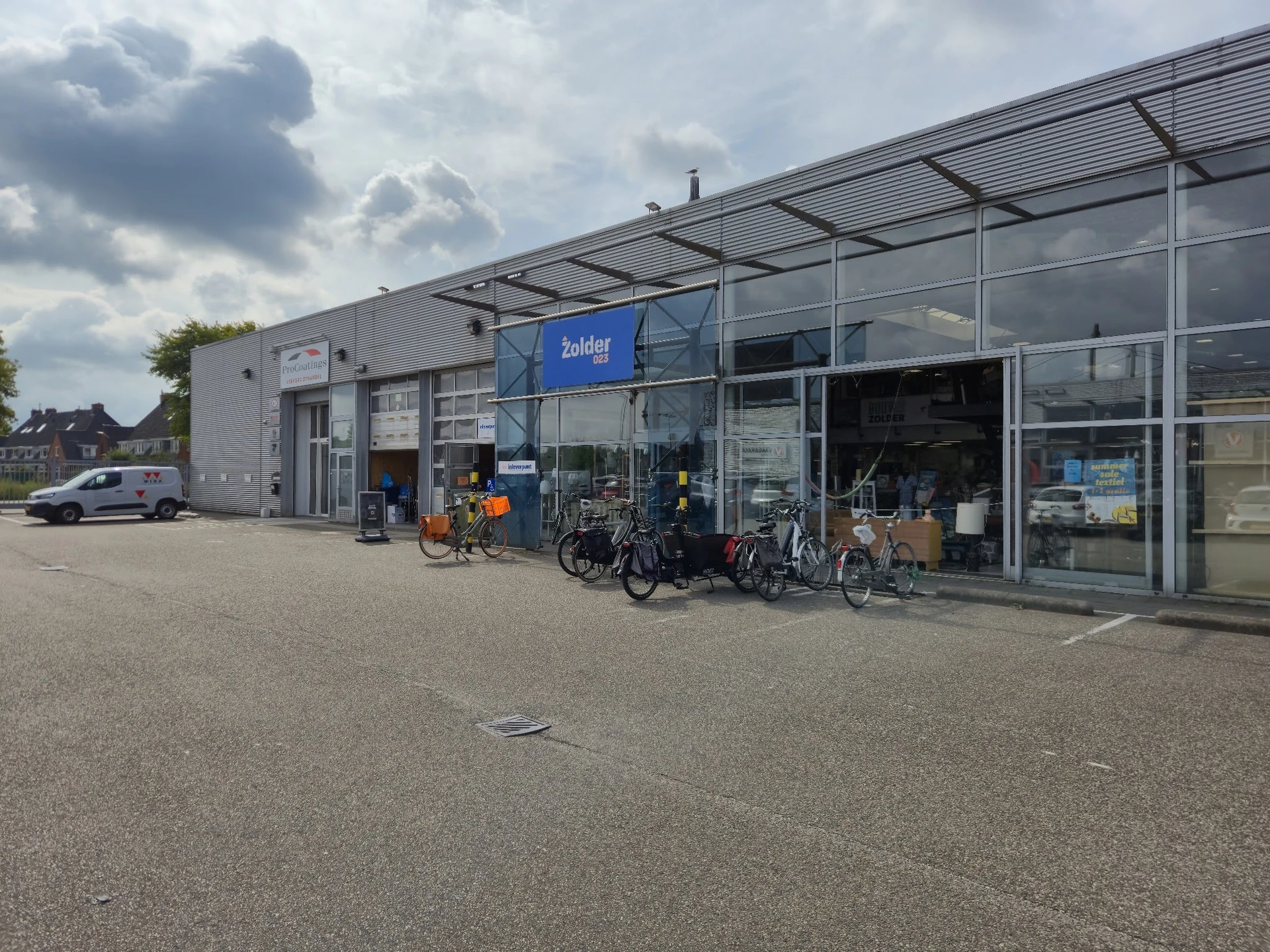 Voorkant kringloopwinkel met glazen pui, blauwe logo, fietsen geparkeerd voor de ingang en parkeerplaats