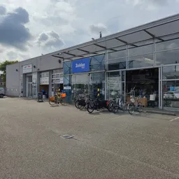 Voorkant kringloopwinkel met glazen pui, blauwe logo, fietsen geparkeerd voor de ingang en parkeerplaats