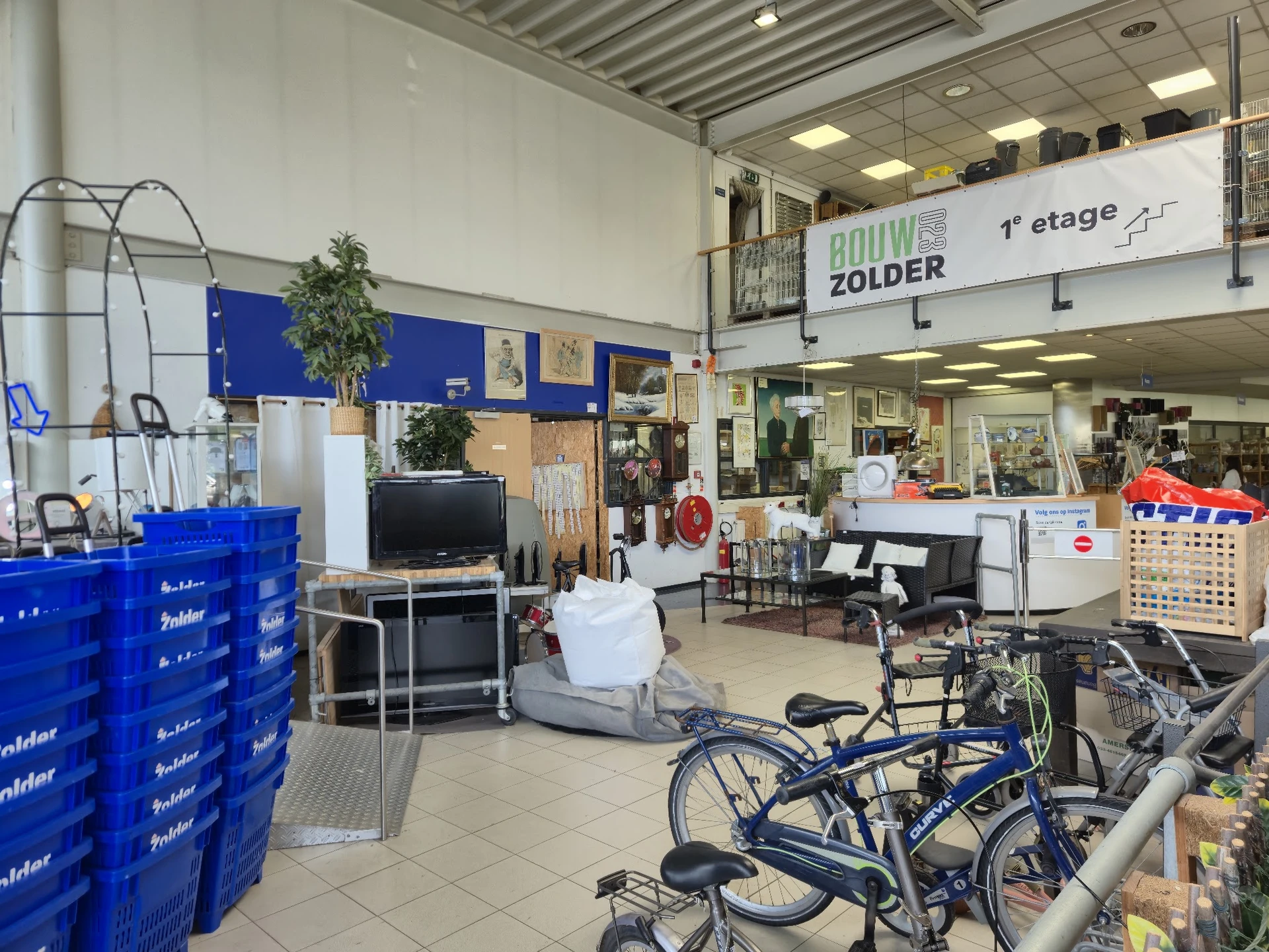 Winkelruimte met fietsenaanbod op voorgrond, blauwe kunststof kratten, stellingen met huishoudwaar en decoratie, trap naar ee