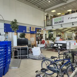 Winkelruimte met fietsenaanbod op voorgrond, blauwe kunststof kratten, stellingen met huishoudwaar en decoratie, trap naar ee