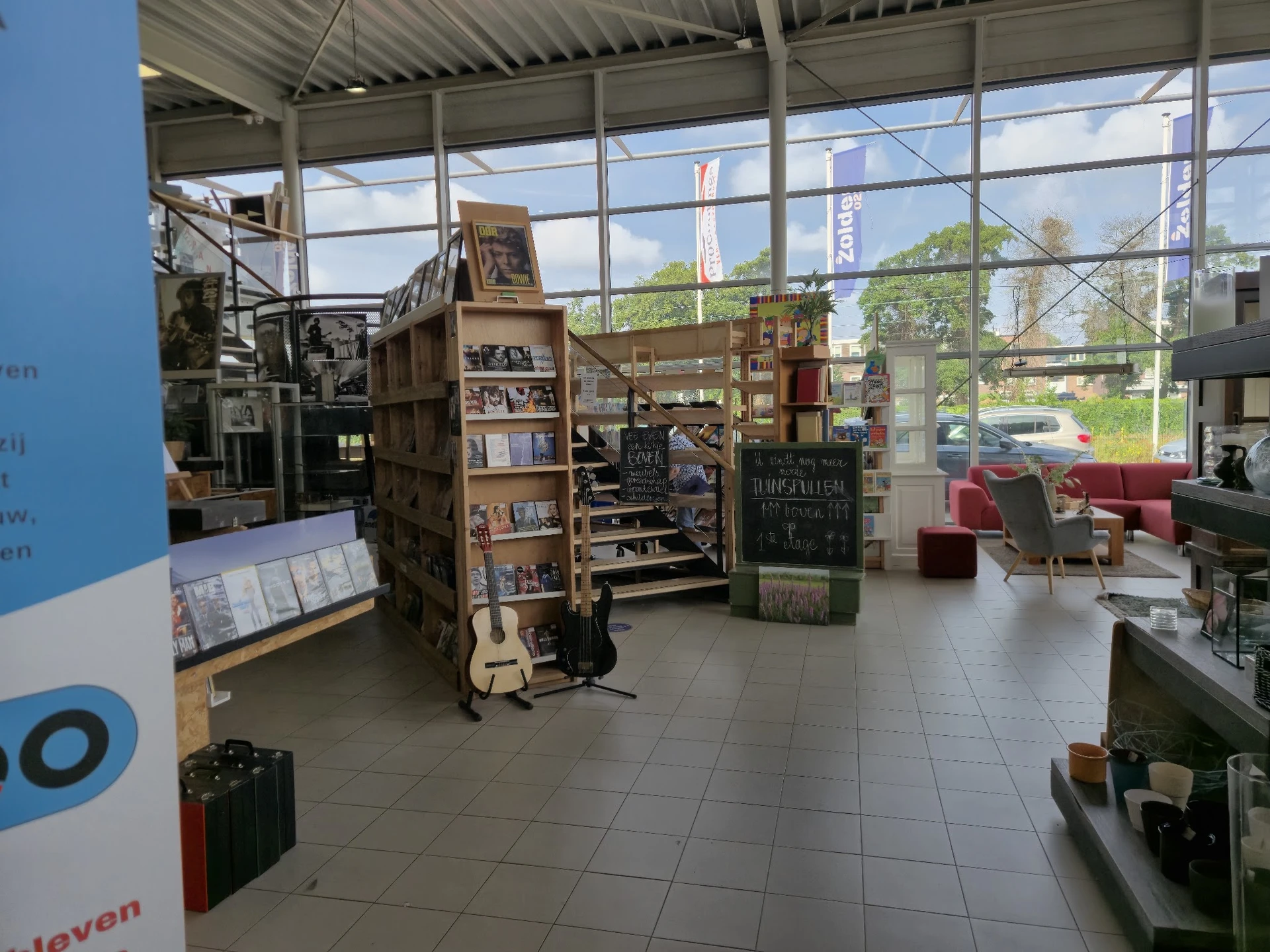 Ruim interieur van kringloopwinkel met stellingen, meubels en spullen verspreid over de vloer, grote ramen met uitzicht op st