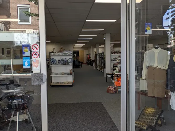 Winkelinterieur met verlichte plafondpanelen, stellingen met spullen, mannequin met kleding rechts, en ingang met glazen deur