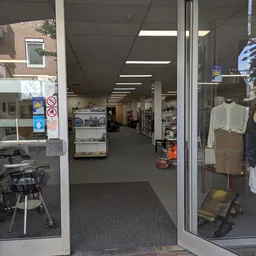 Winkelinterieur met verlichte plafondpanelen, stellingen met spullen, mannequin met kleding rechts, en ingang met glazen deur